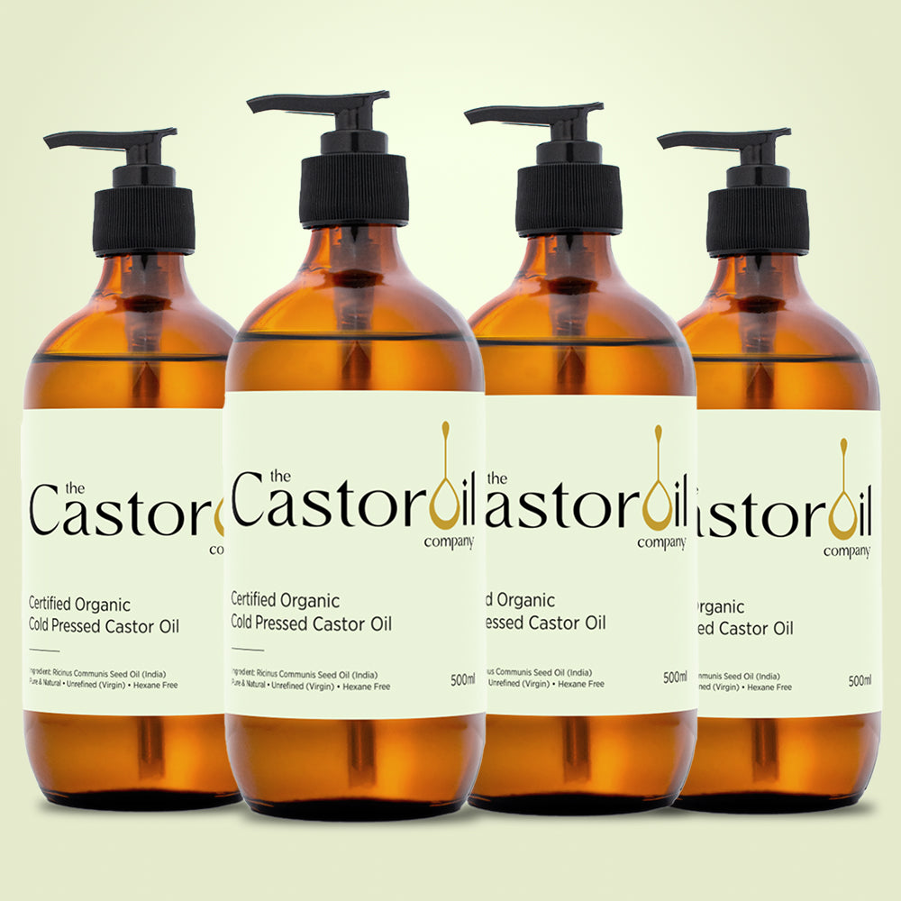 Four amber glass pump bottles of The Castor Oil Company's Certified Organic Cold Pressed Castor Oil arranged in a row against a pale yellow background.